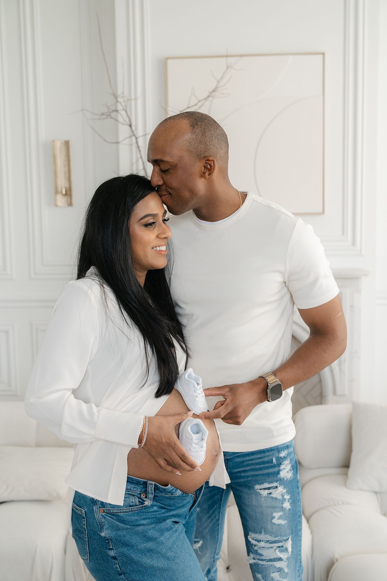 Happy couple holding baby shoes near pregnant belly in bright white modern room.