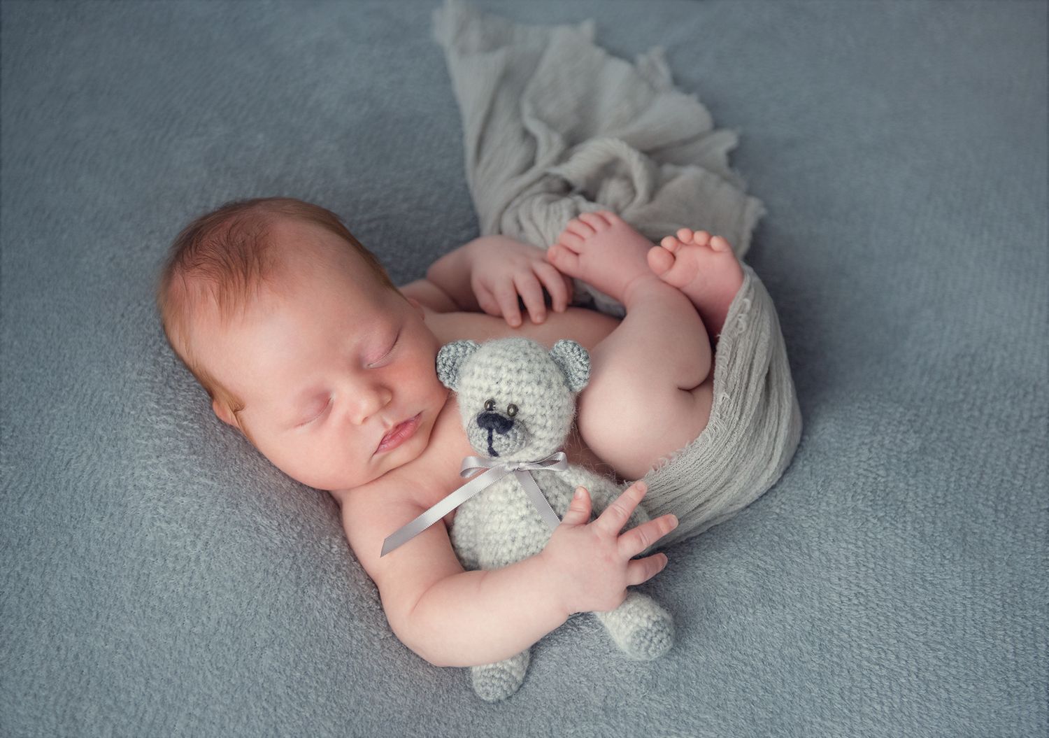 Sleeping newborn baby holding a small gray crocheted teddy bear on a soft gray blanket.