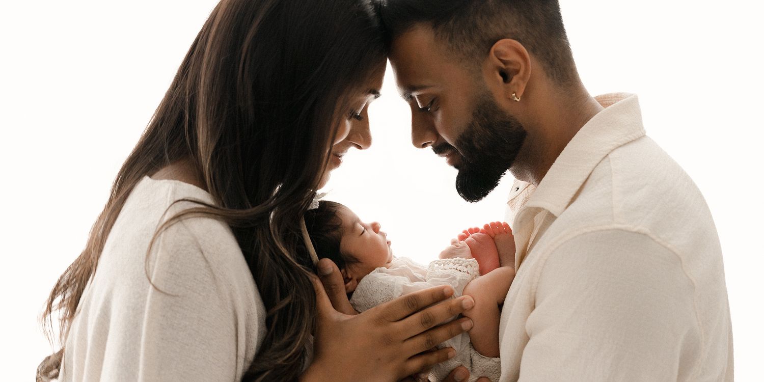 A couple in white outfits tenderly hold their newborn baby together against a white background.