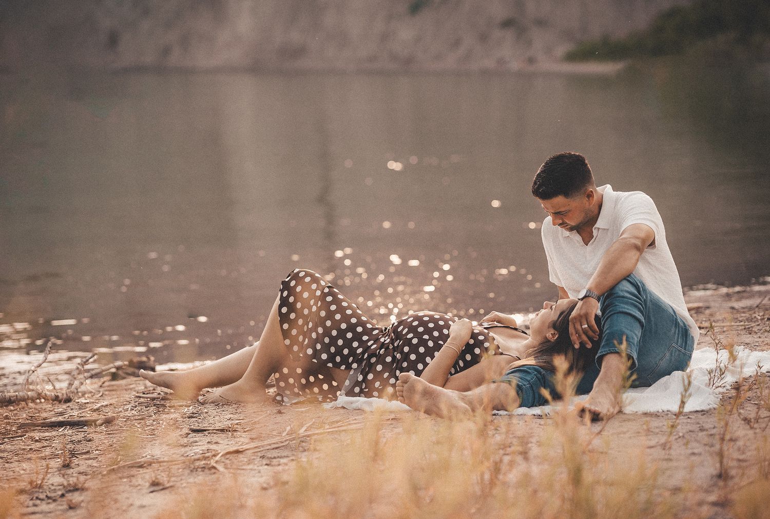 Expecting couple sharing romantic moment by lakeside, woman in polka dot skirt lying on mans lap.