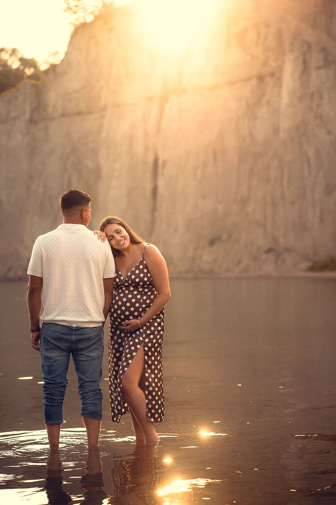 Expecting couple standing in shallow water at golden sunset, woman in polka dot maternity dress.