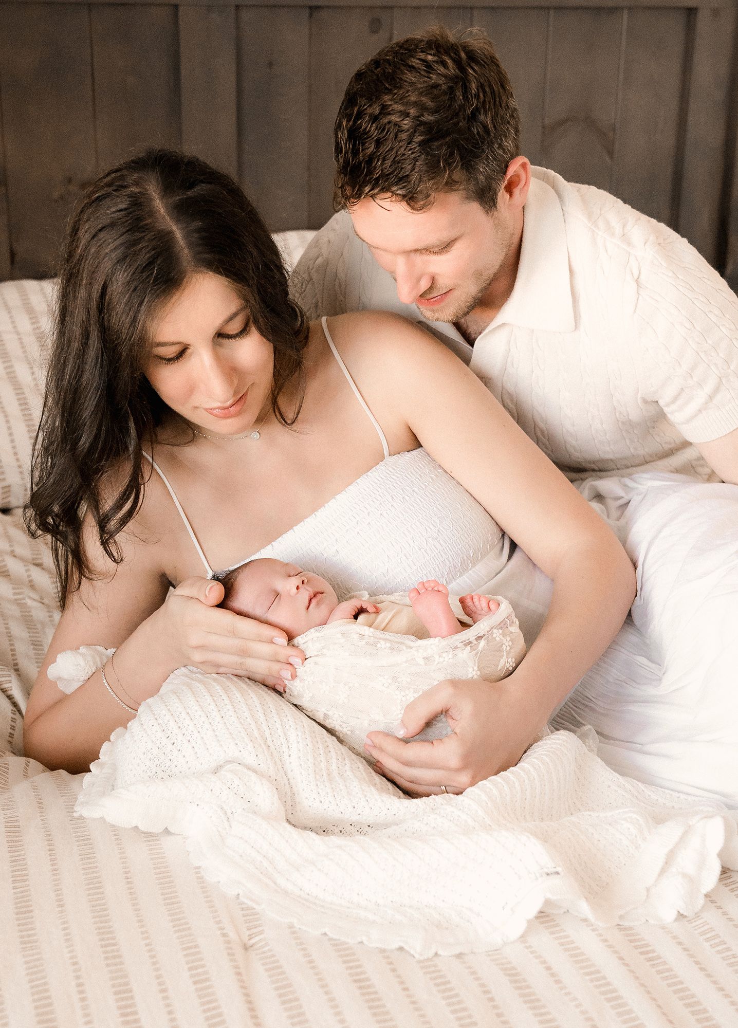 Smiling new parents cuddle their sleeping newborn baby wrapped in a soft white knit blanket while resting together on a bed.