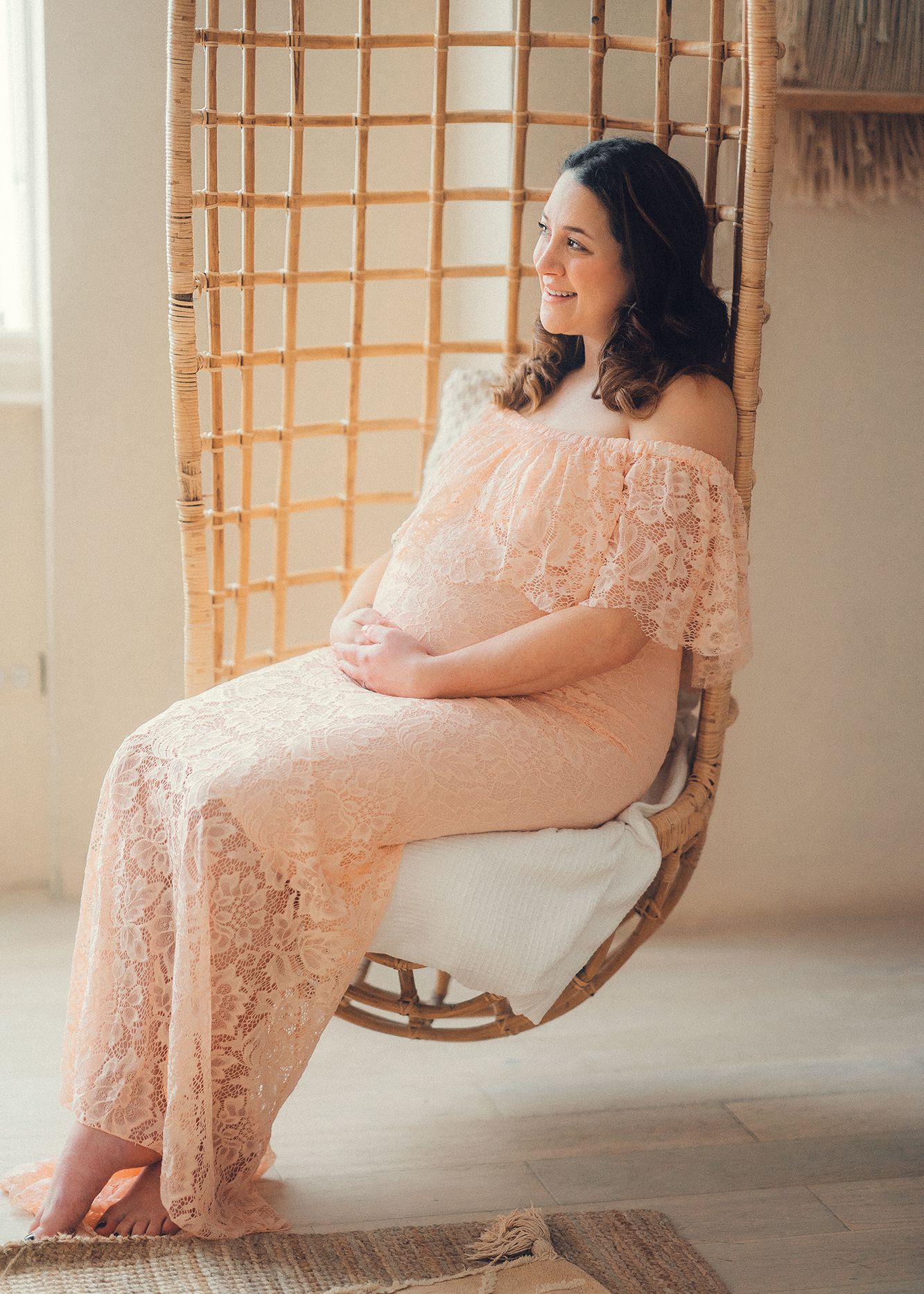 Smiling pregnant woman in pink lace maternity outfit sitting in a rattan hanging chair.