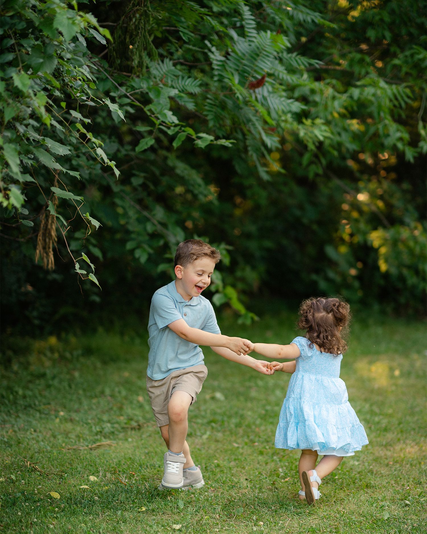 Two young children laugh and spin together holding hands on a lush green lawn under tall trees.