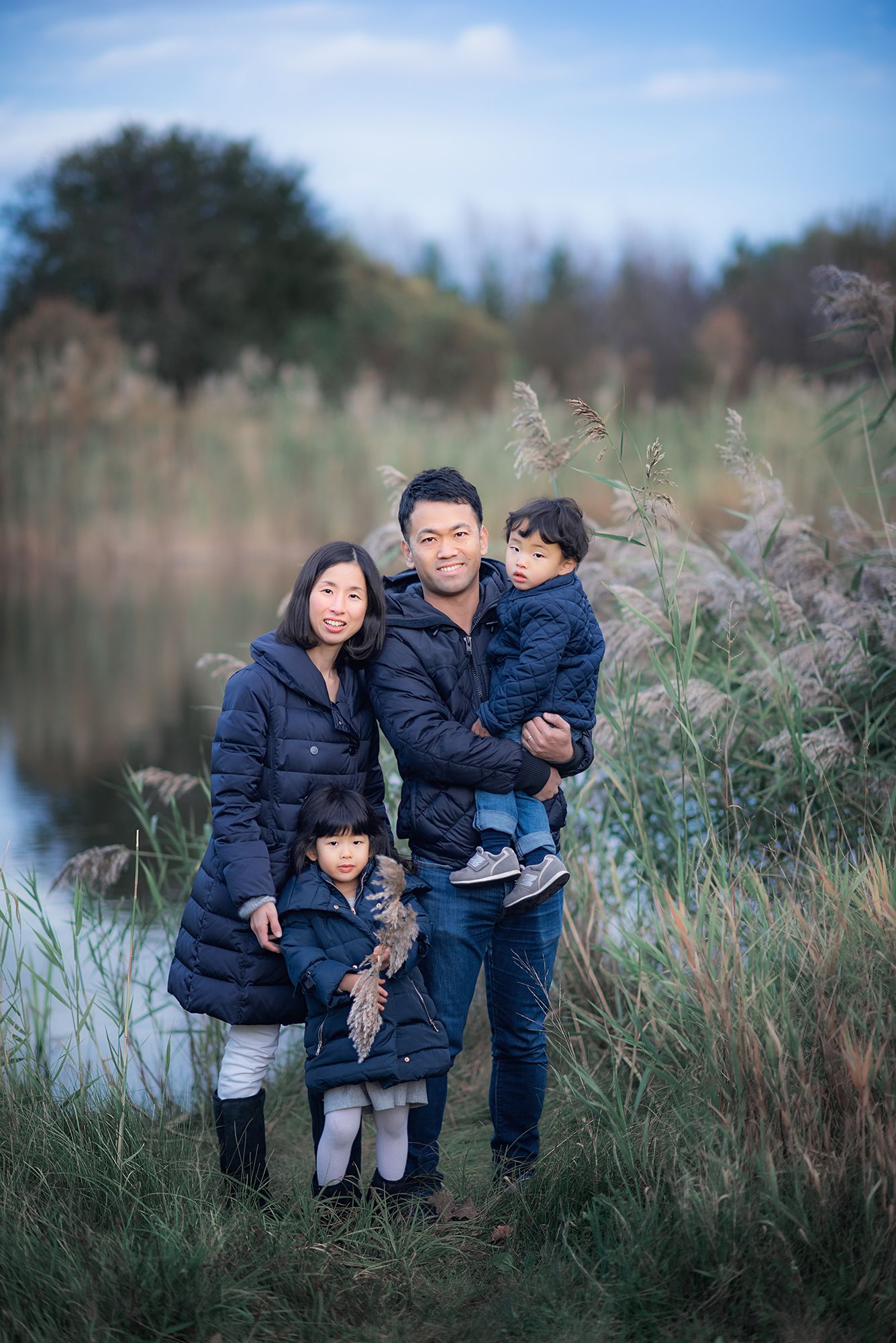 An Asian family of four in matching navy jackets poses near a serene lake surrounded by tall grass.