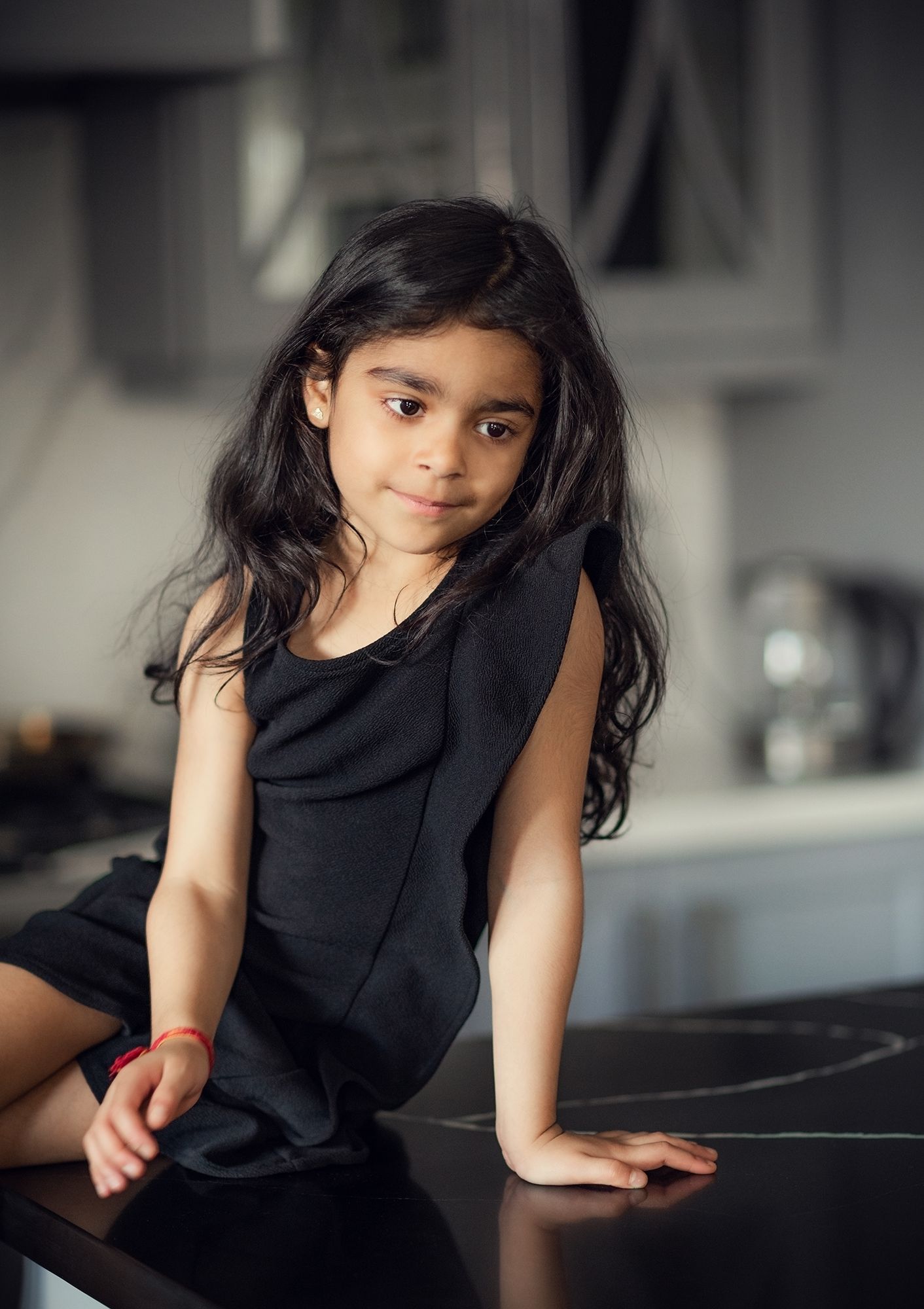 Young girl with long dark hair in black dress sitting on kitchen counter smiling.