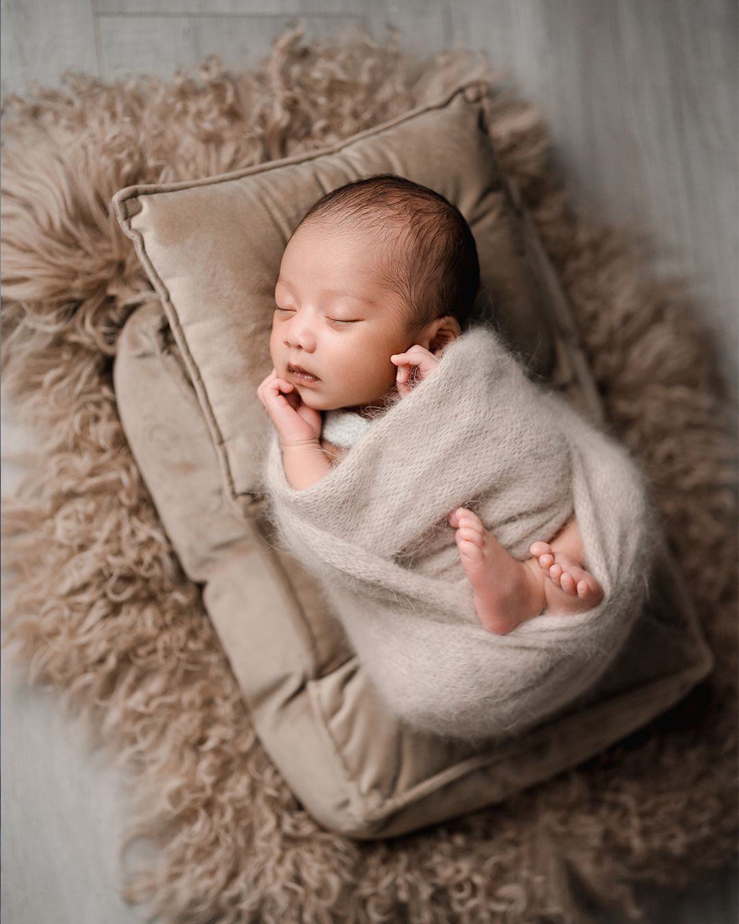 Peacefully sleeping newborn wrapped in beige knit swaddle on velvet pillow with fluffy rug.
