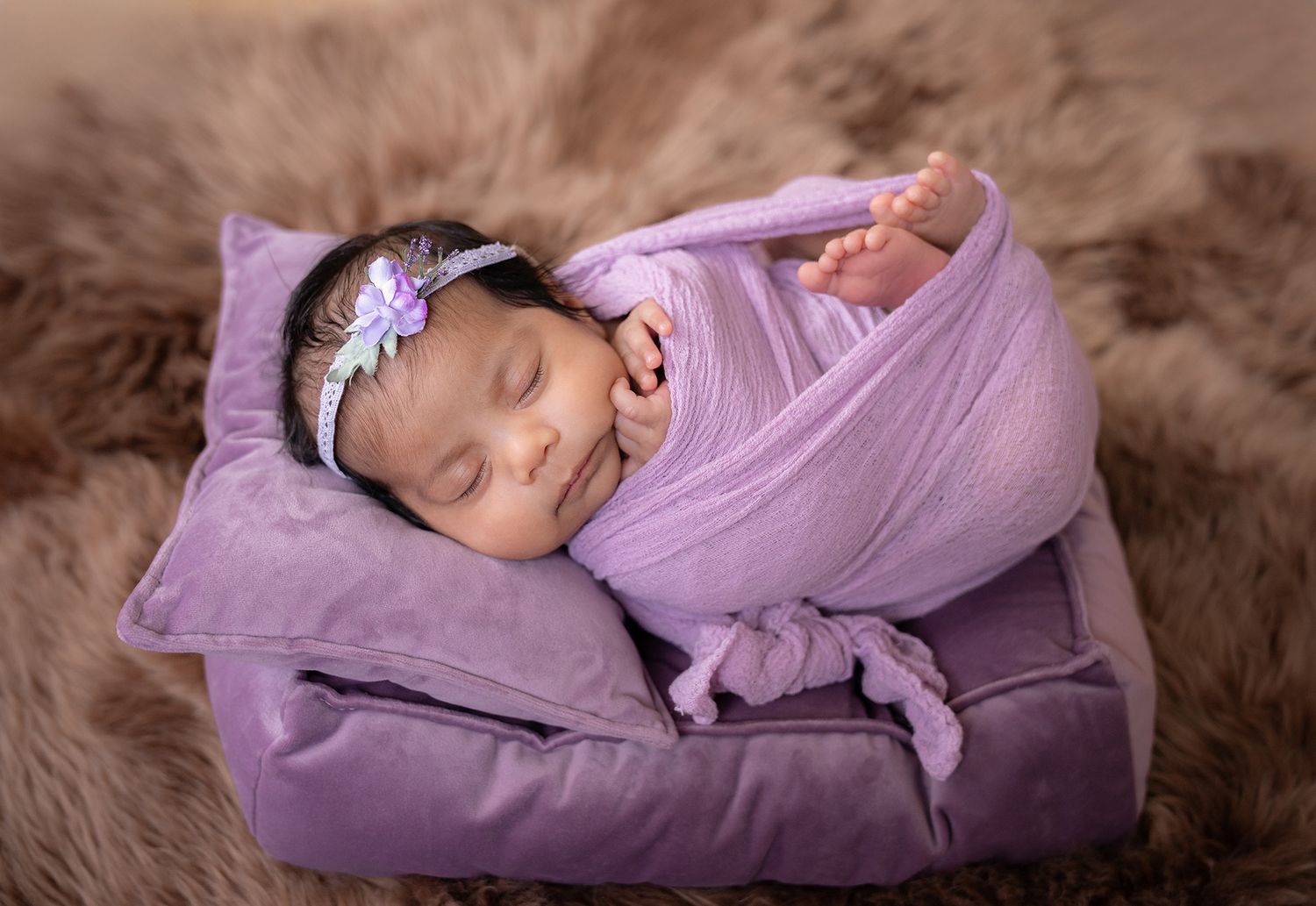 Newborn baby girl swaddled in purple wrap with floral headband sleeping on a velvet pillow.