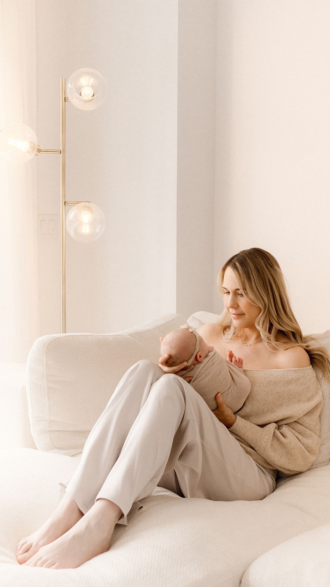 Mother in a beige off-shoulder sweater sits barefoot on a cream sofa, cradling her newborn baby in a bright, minimalist living room.