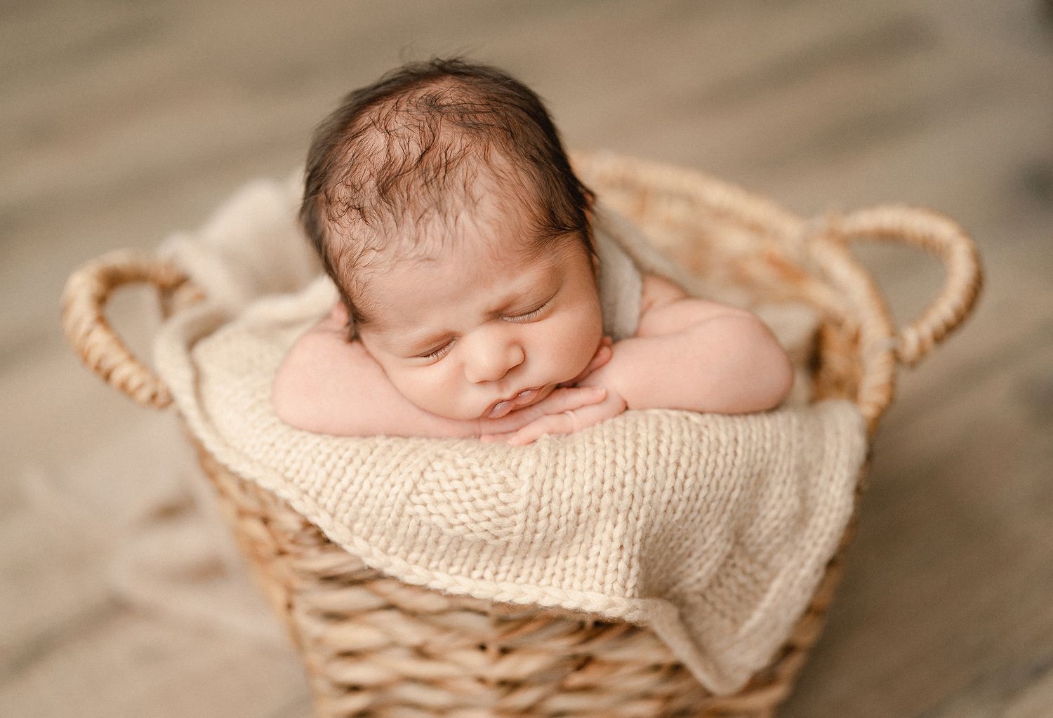Sleeping newborn baby resting in a woven wicker basket lined with a cream knit blanket during a soft newborn photoshoot.