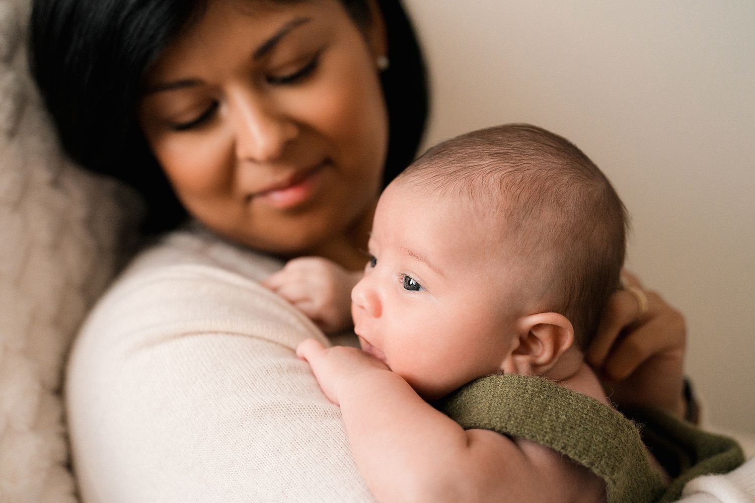 A smiling mother with dark hair gently holds her wide-eyed newborn baby against her shoulder in a warm, softly lit indoor setting.
