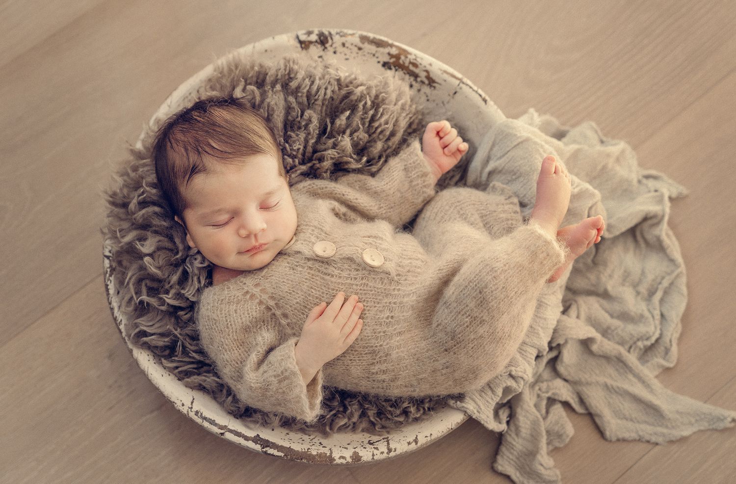 Sleeping newborn baby in knit gray outfit curled in a round wooden bowl with fur.