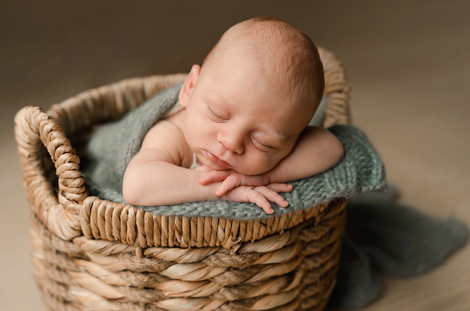 Peaceful newborn baby sleeping curled up in a woven wicker basket with teal fabric.