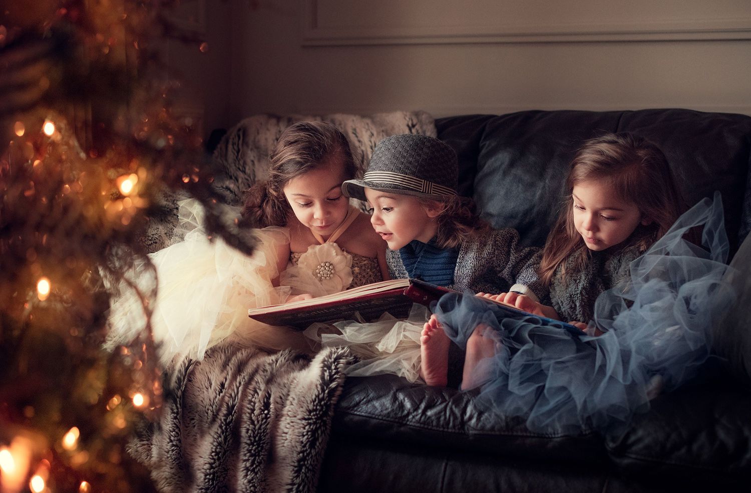 Three children reading a glowing book together on couch by Christmas tree lights.