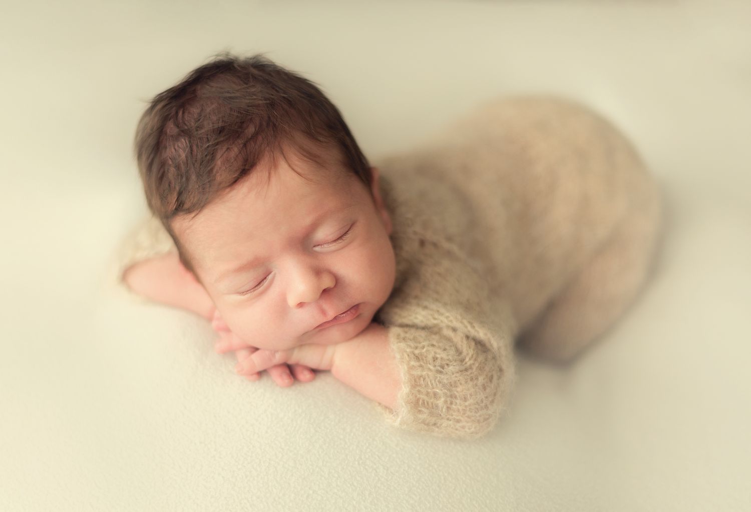 Peaceful newborn baby sleeping in a beige knit romper on a soft white blanket.