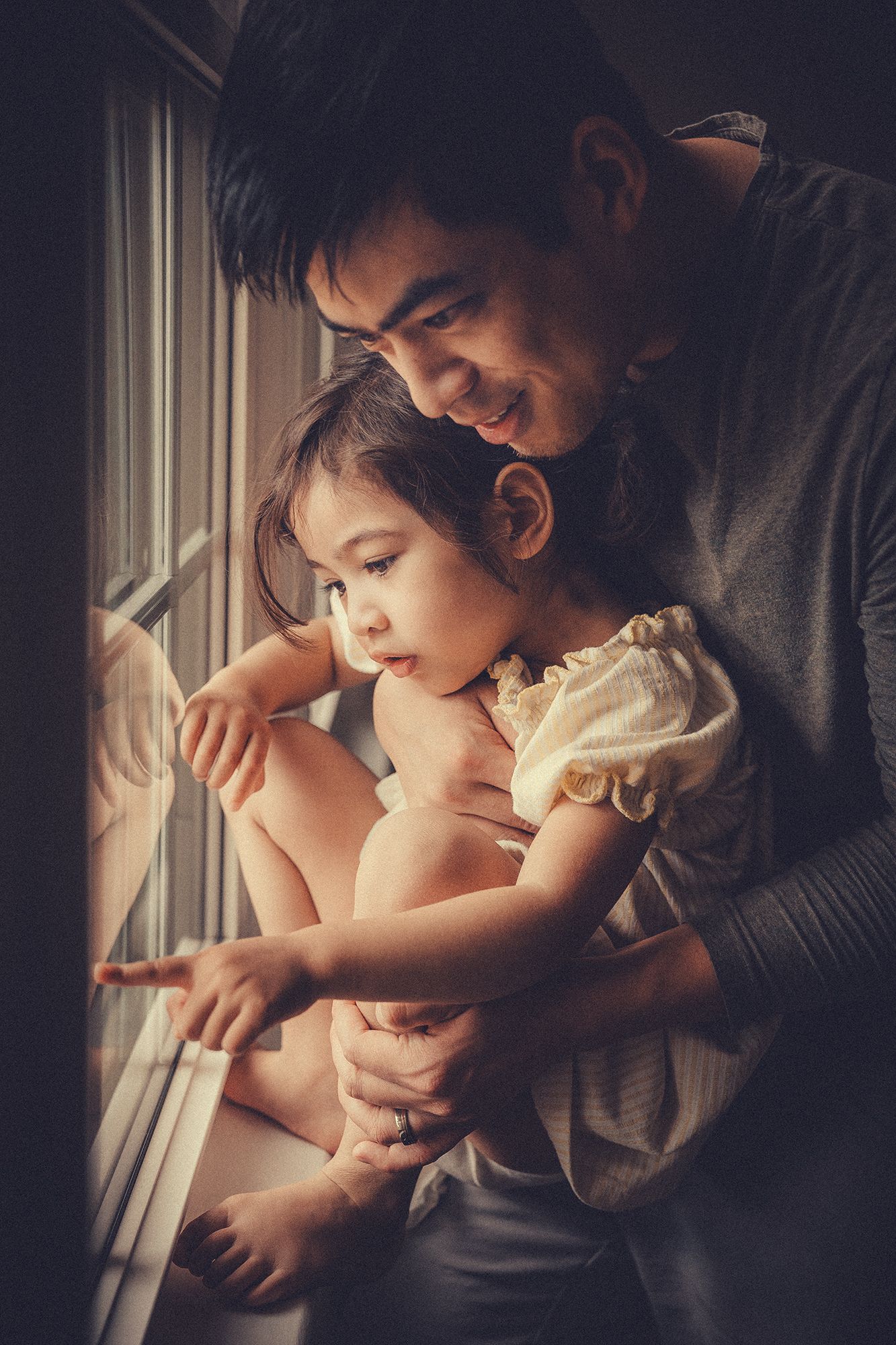 A smiling father holds his young daughter as she points at something through a window.