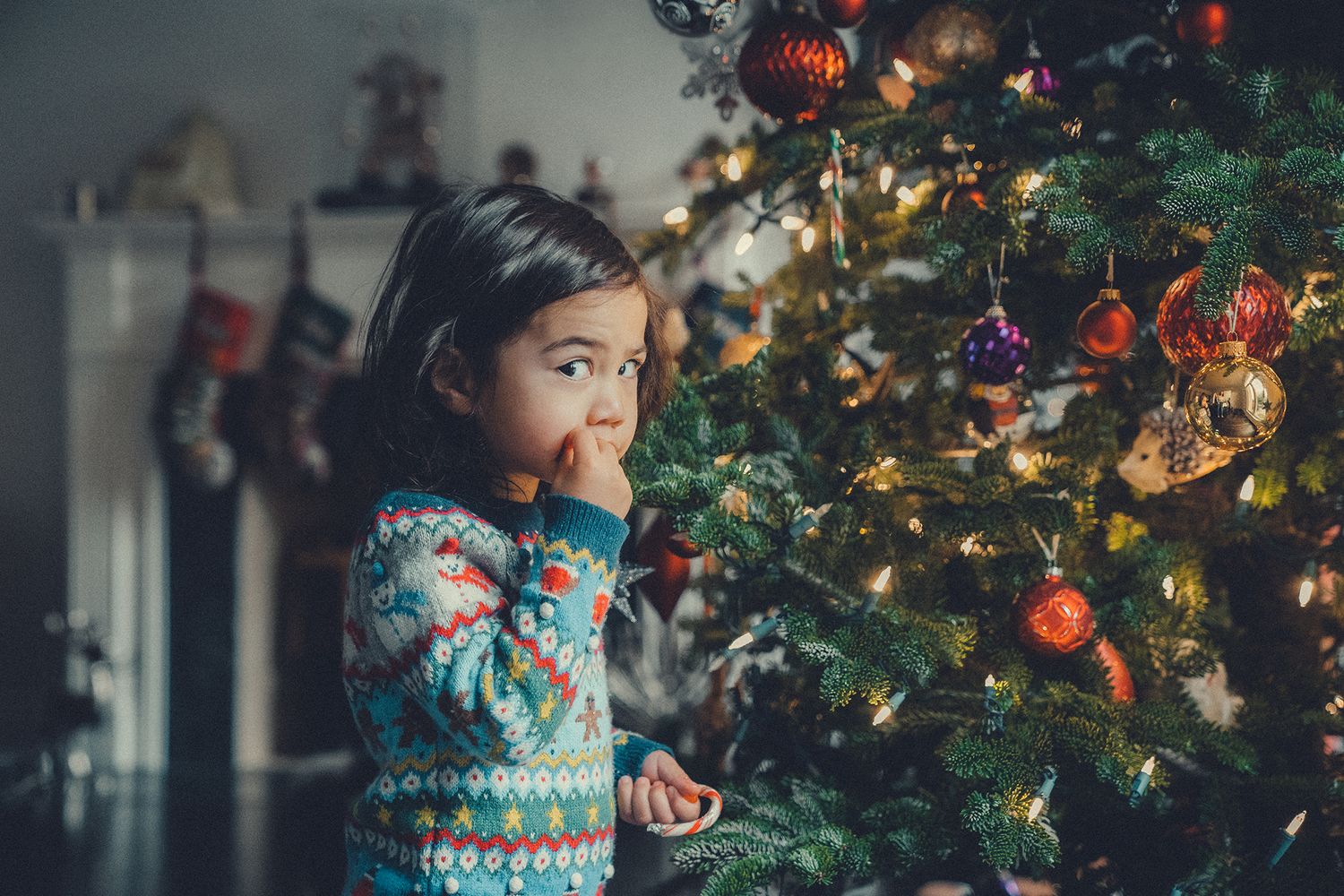 Curious toddler in festive sweater admiring decorated Christmas tree with colorful ornaments indoors.