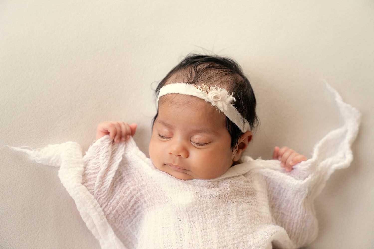 Newborn baby girl with dark hair wearing a white floral headband wrapped in white gauze fabric.