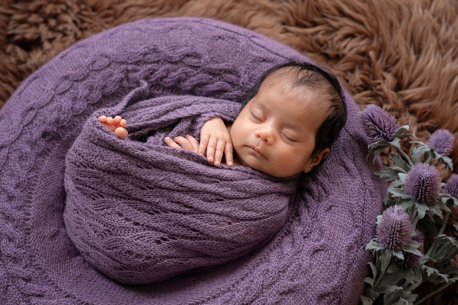 Sleeping newborn baby swaddled in purple knit wrap in a bowl with purple flowers.
