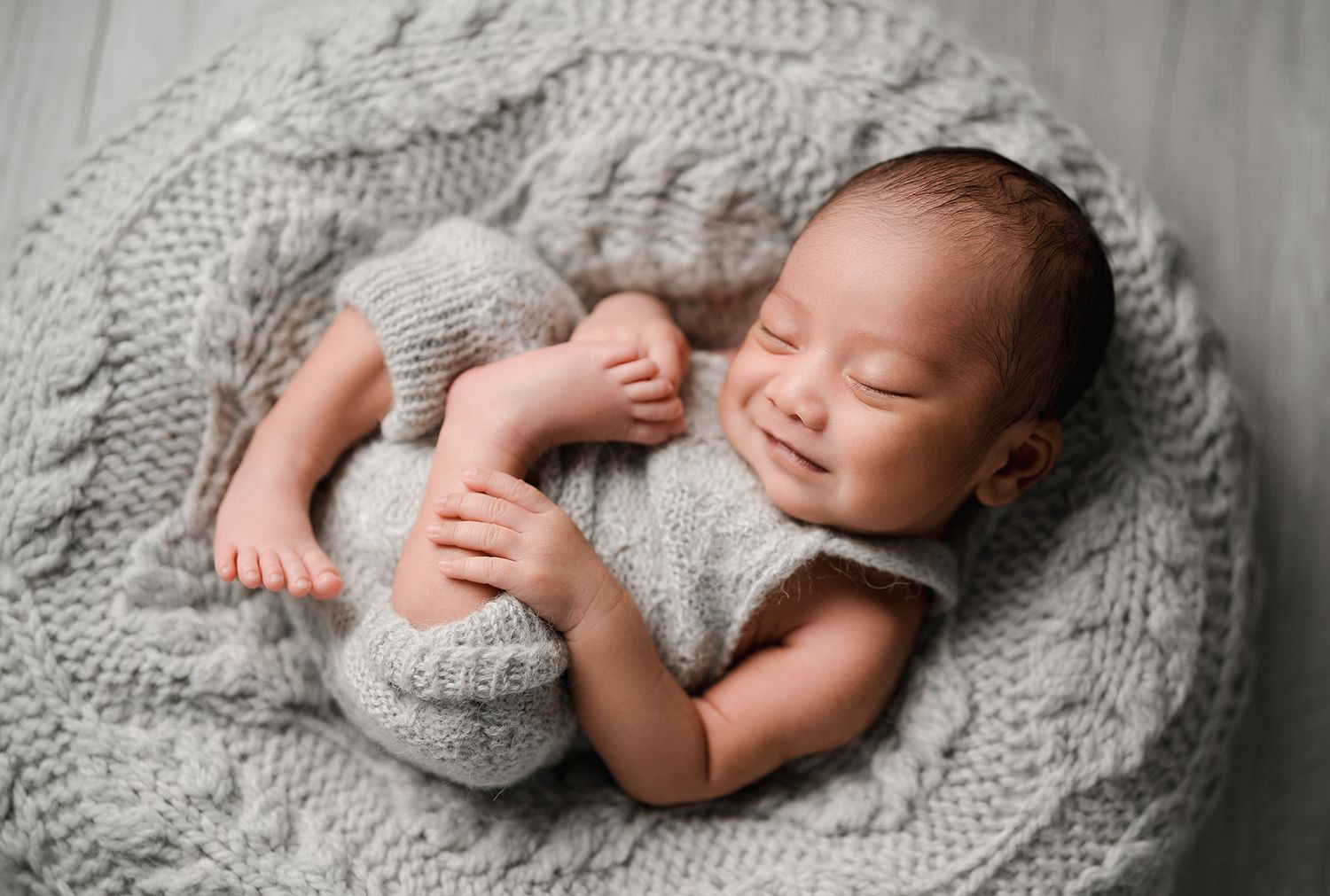 Smiling newborn baby in a gray knit romper sleeping curled up on a cozy cable-knit blanket in a round basket.