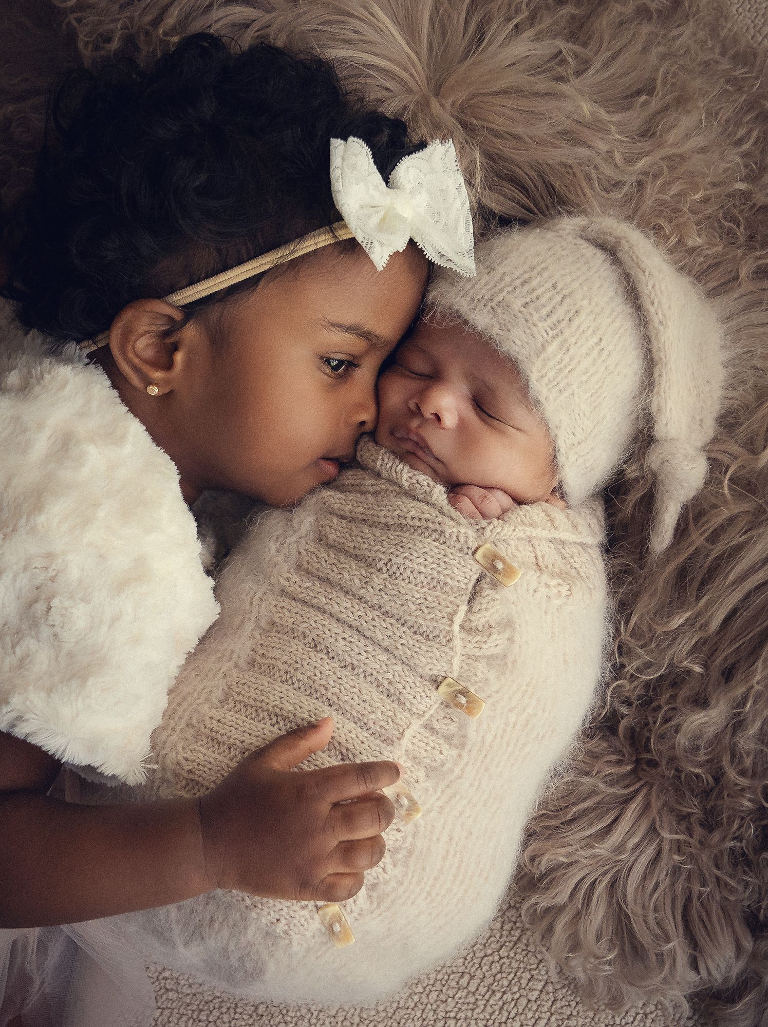 Young girl tenderly kissing a sleeping newborn baby swaddled in a cream knit wrap.