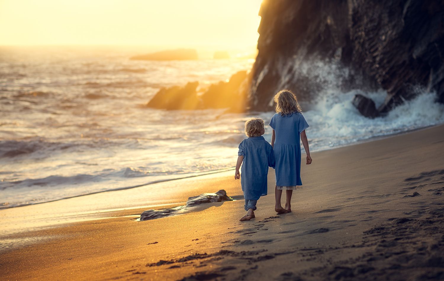 Two young children in blue outfits walk hand in hand along a sandy beach at golden sunset.