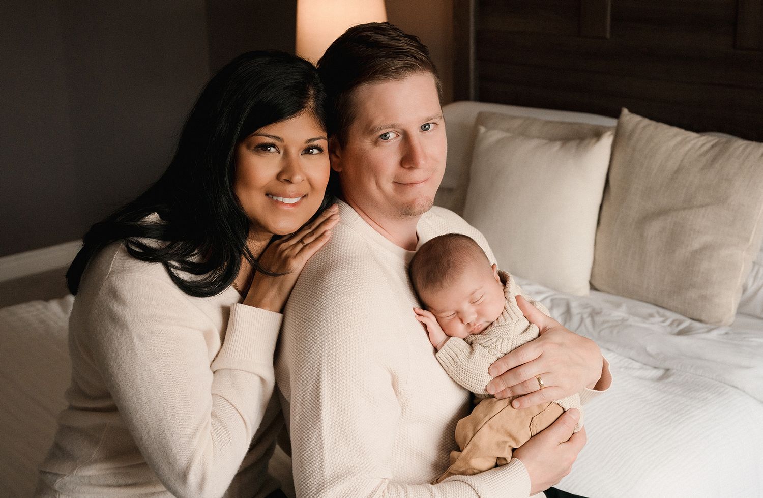 A smiling woman and a man in cream sweaters sit close on a bed, lovingly holding their sleeping newborn baby between them.