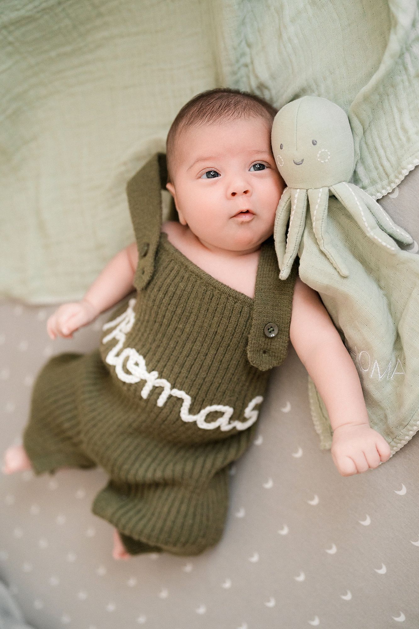Newborn baby in an olive green knit romper with the name Thomas stitched in white, lying beside a plush octopus toy on a soft starry sheet.