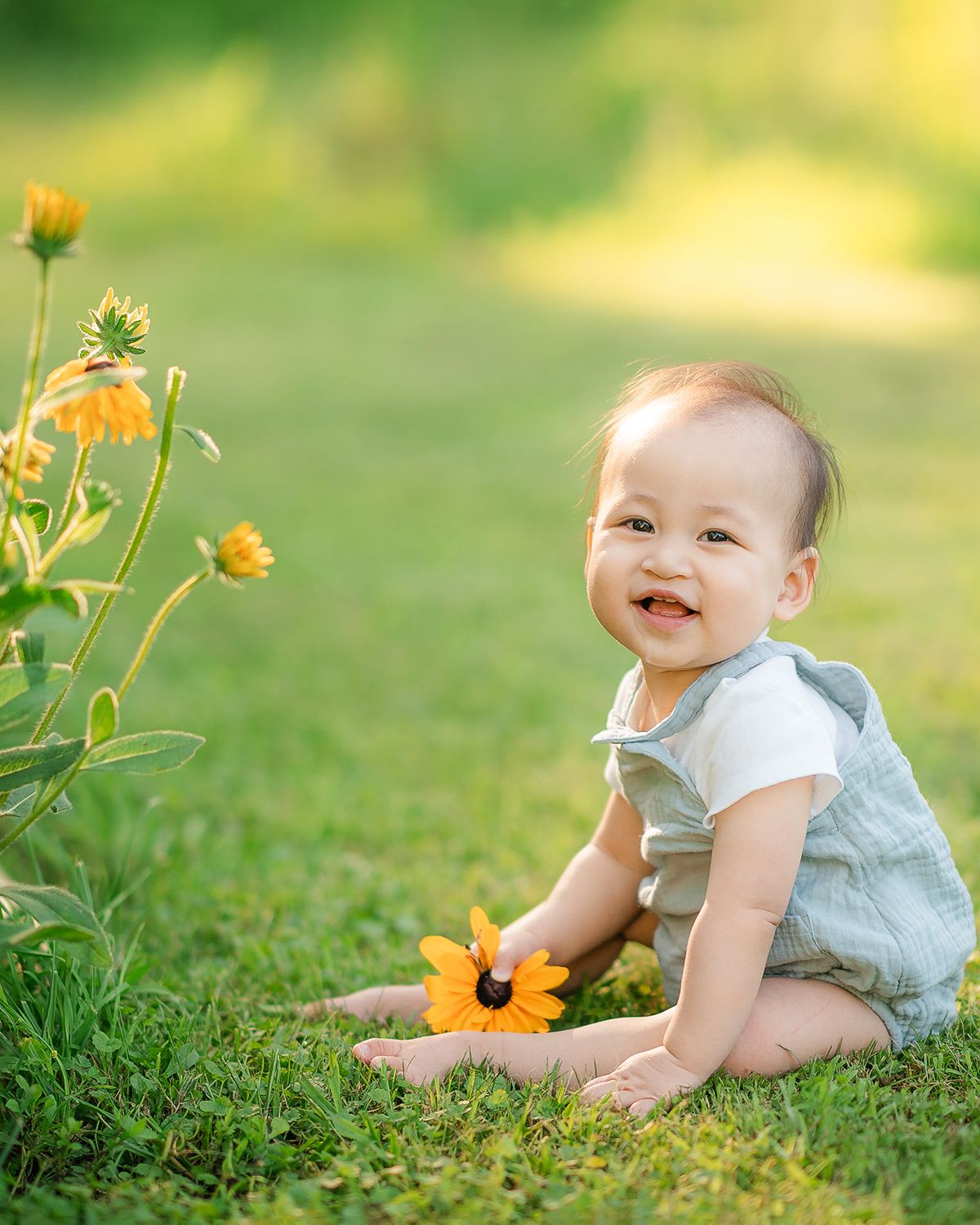 Happy baby sitting on green grass holding a yellow flower in sunny garden.