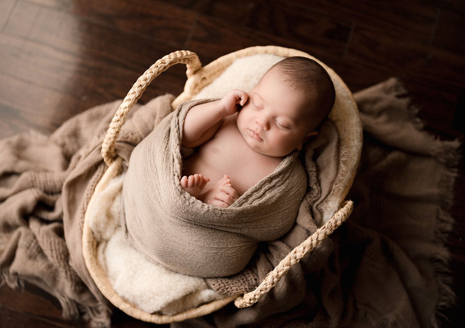 Sleeping newborn baby wrapped in a soft taupe swaddle, resting in a woven basket on a rustic wooden floor with a knit blanket.