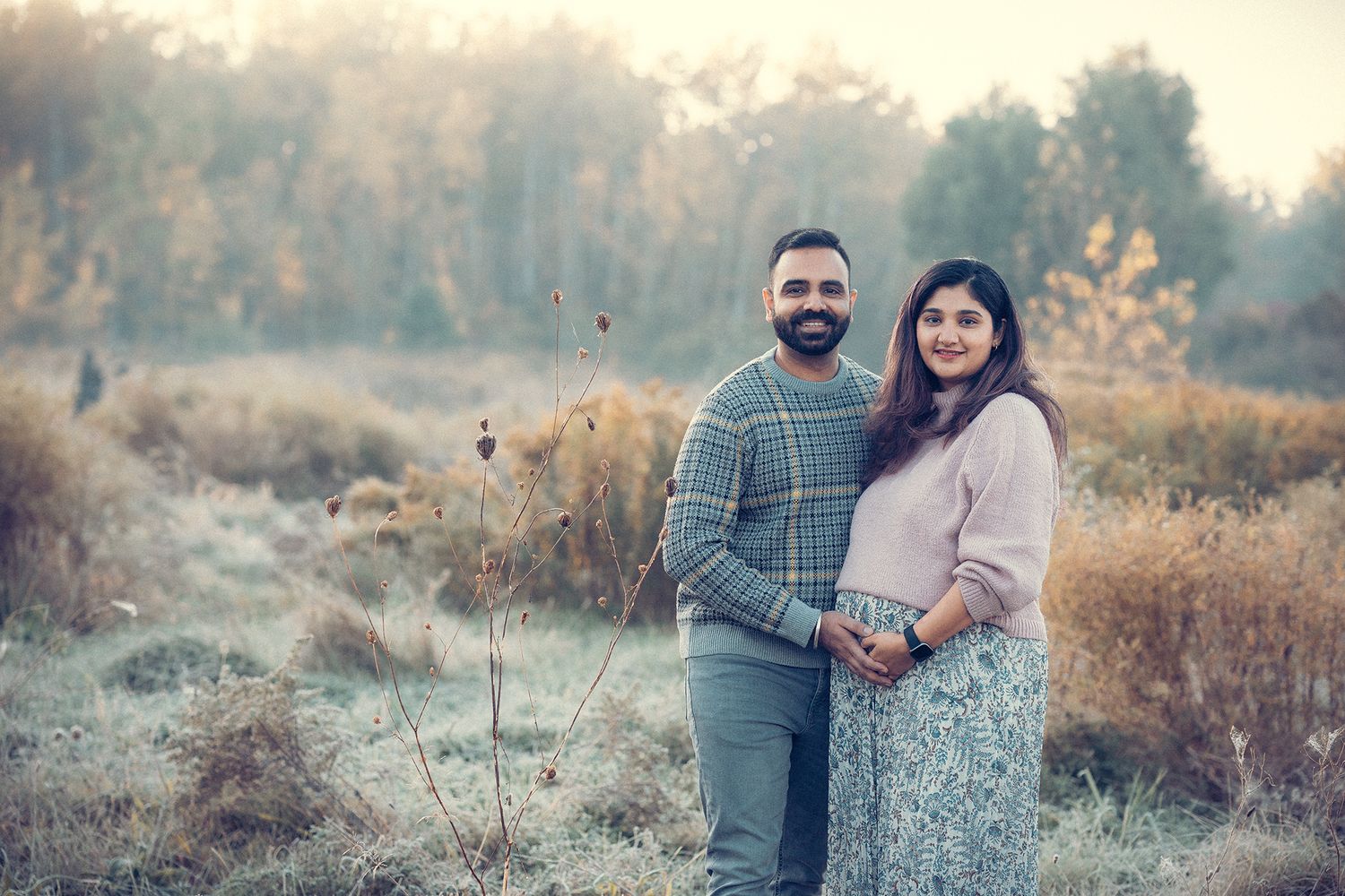 Couple holding baby bump together in frosty autumn field at sunrise.