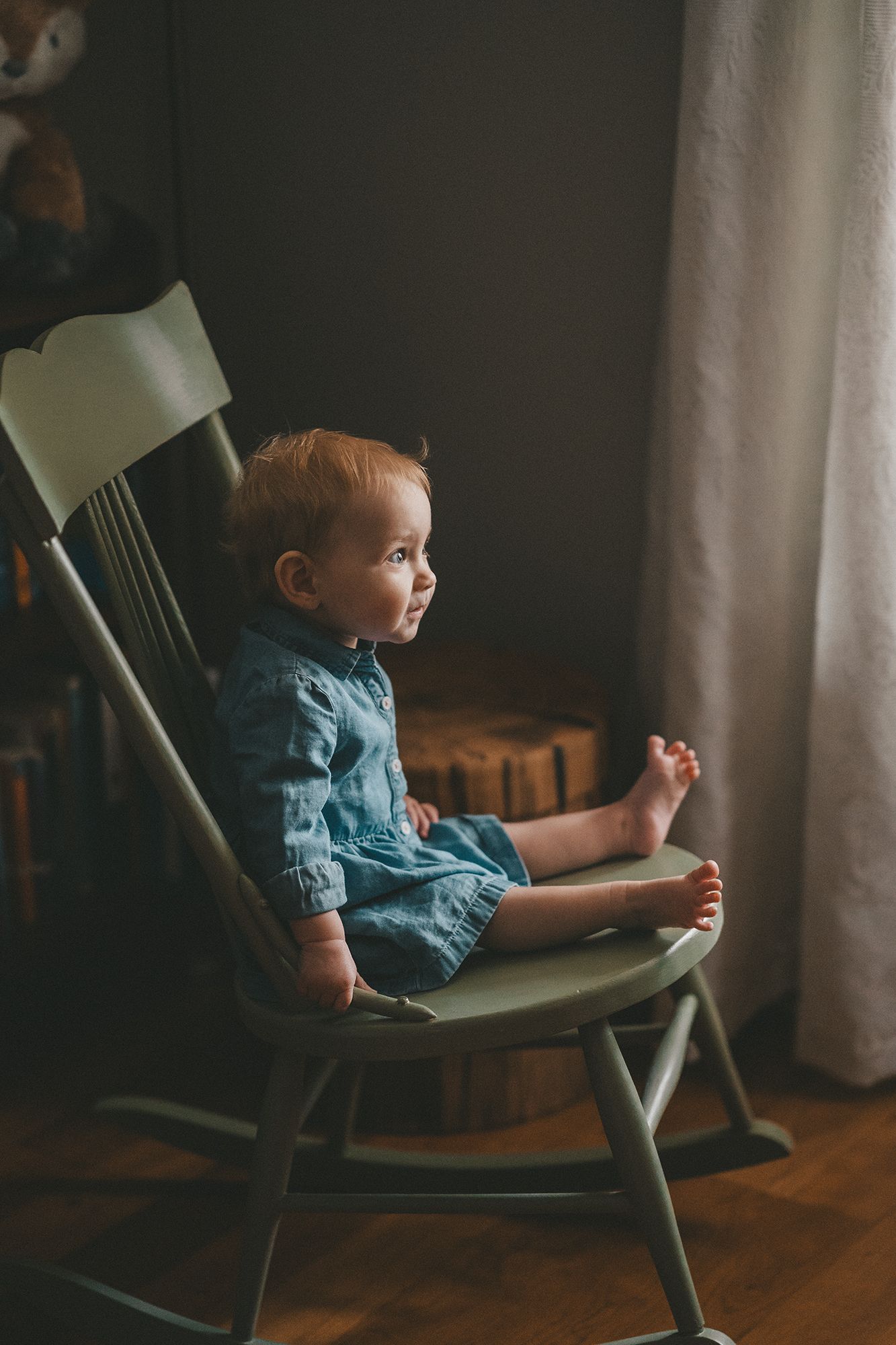 Adorable baby in denim outfit sitting in wooden rocking chair looking toward window indoors.