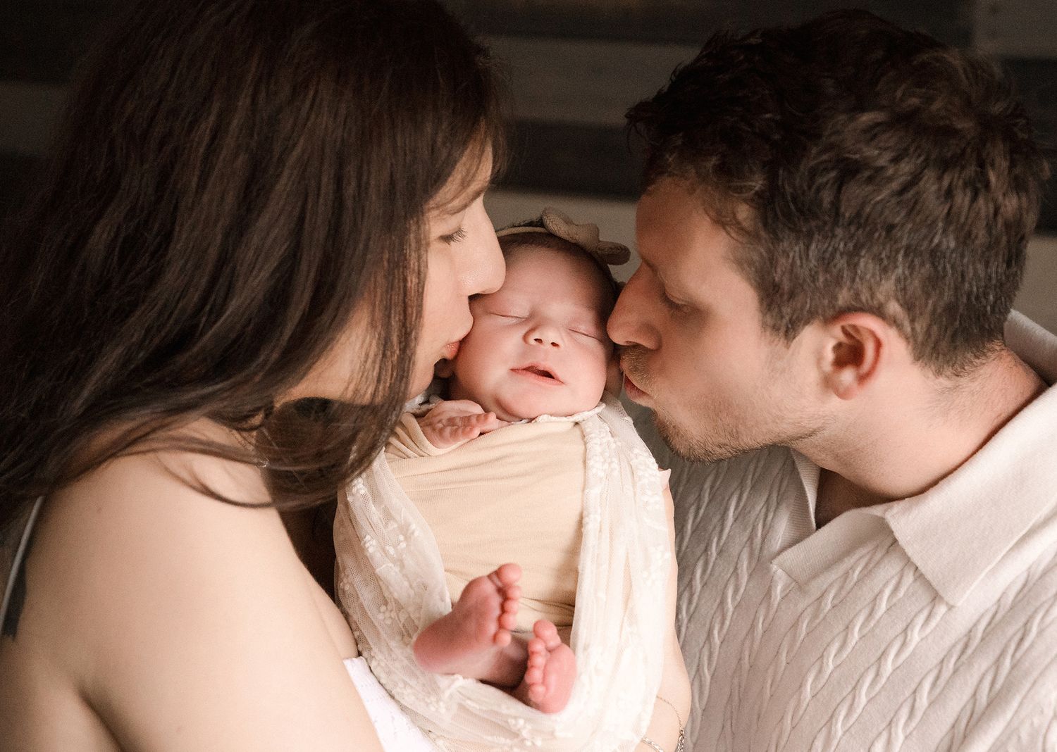 A mother and father gently kiss their sleeping newborn baby's cheeks while the infant rests wrapped in a soft cream blanket.
