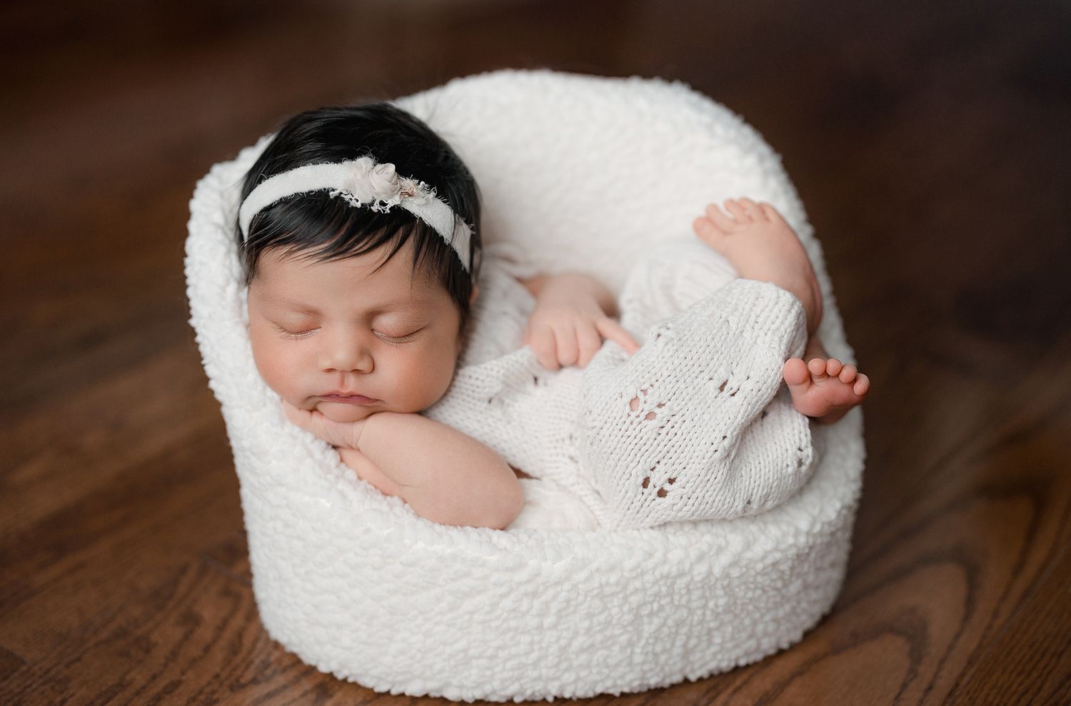 A peaceful newborn baby sleeps curled up in a tiny white fluffy chair during a photo session.