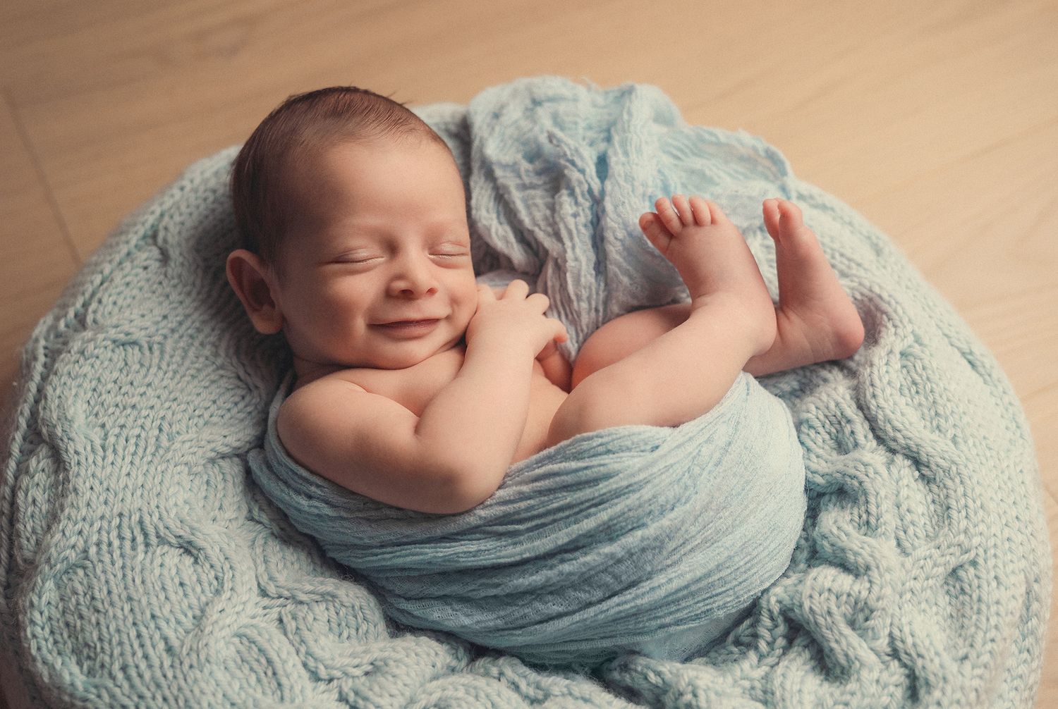 Smiling sleeping newborn wrapped in a light blue knit swaddle posed in a round knit bowl.