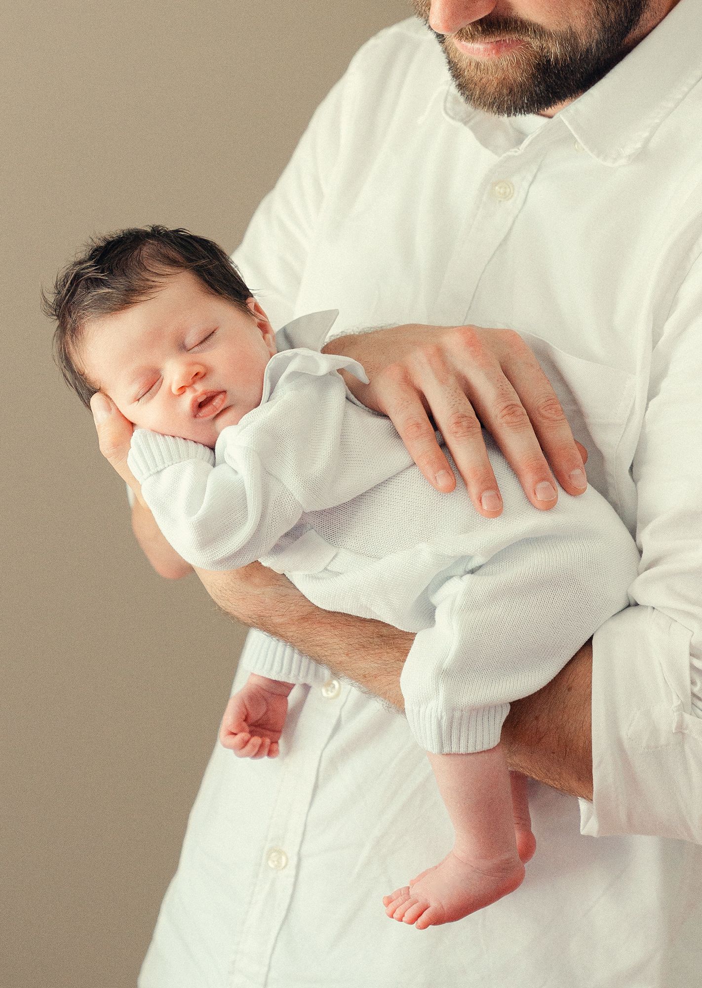 Bearded father in white shirt cradling his sleeping newborn baby dressed in white outfit.