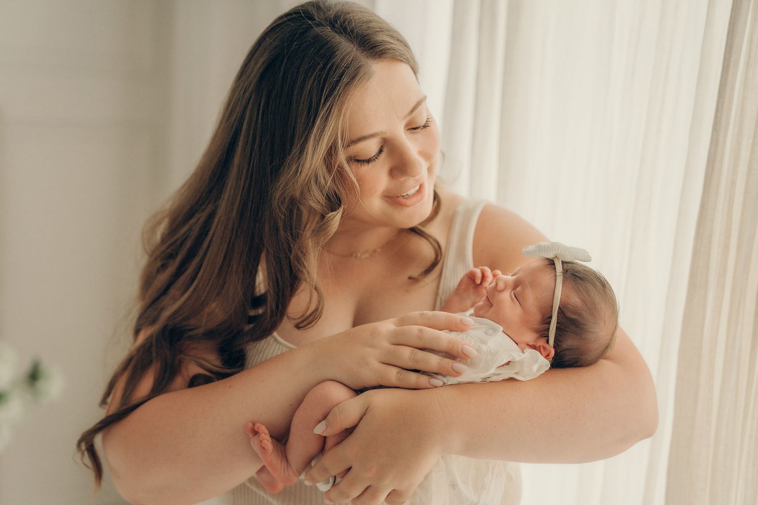 A smiling mother with long hair tenderly holds her sleeping newborn baby near a bright window.