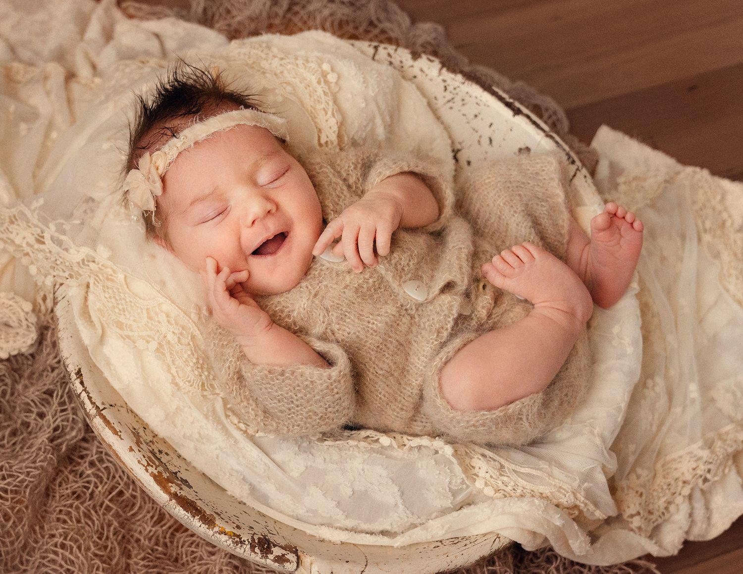 Smiling newborn baby lying in a vintage basket with soft cream lace wraps during a professional photo session.