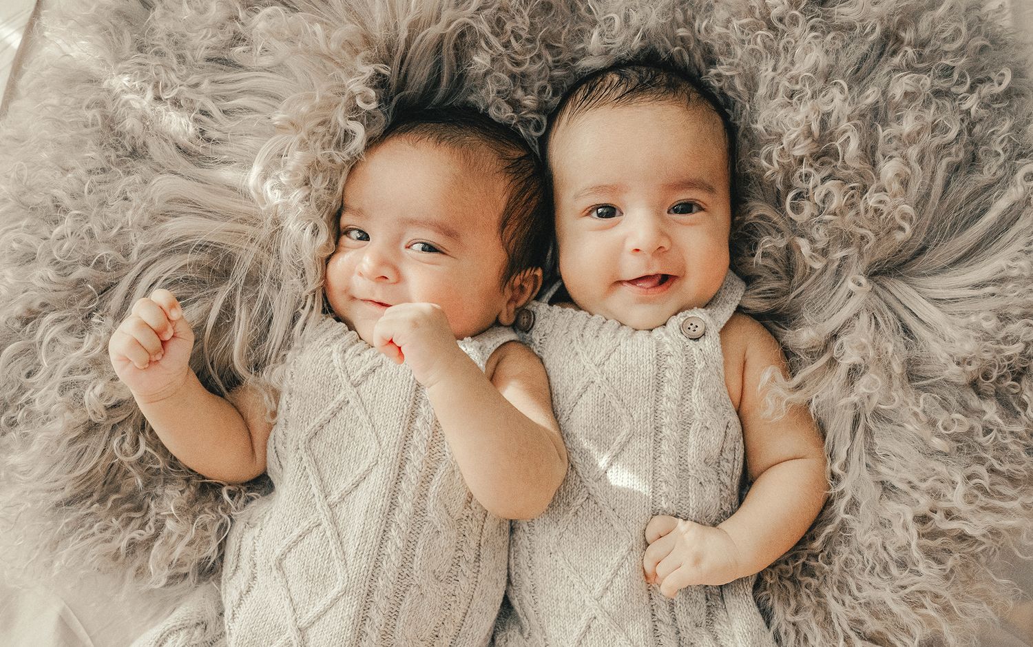 Two adorable smiling twin babies lying on a fluffy gray fur blanket wearing matching gray knit outfits.