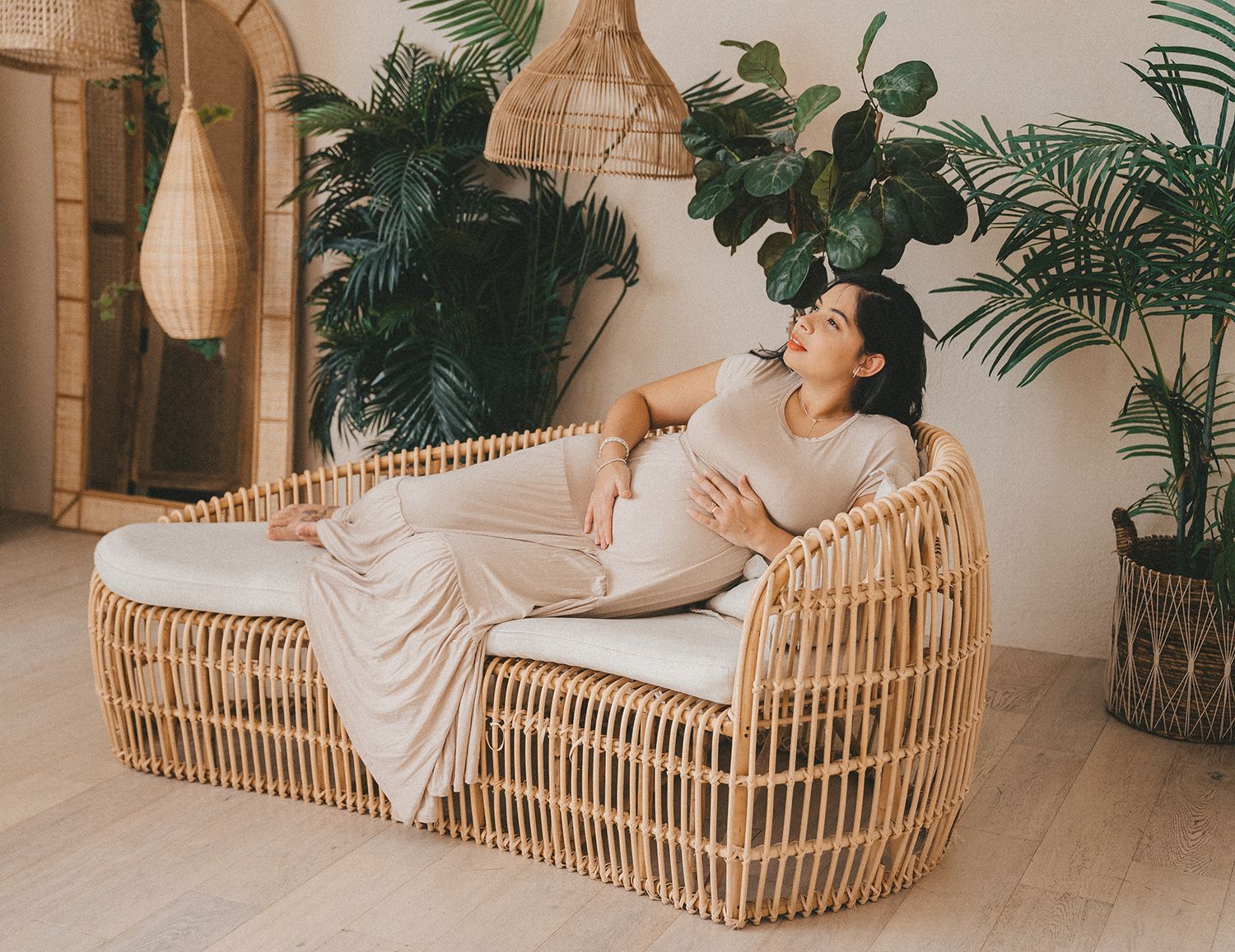 Pregnant woman relaxing on rattan sofa surrounded by tropical indoor plants.