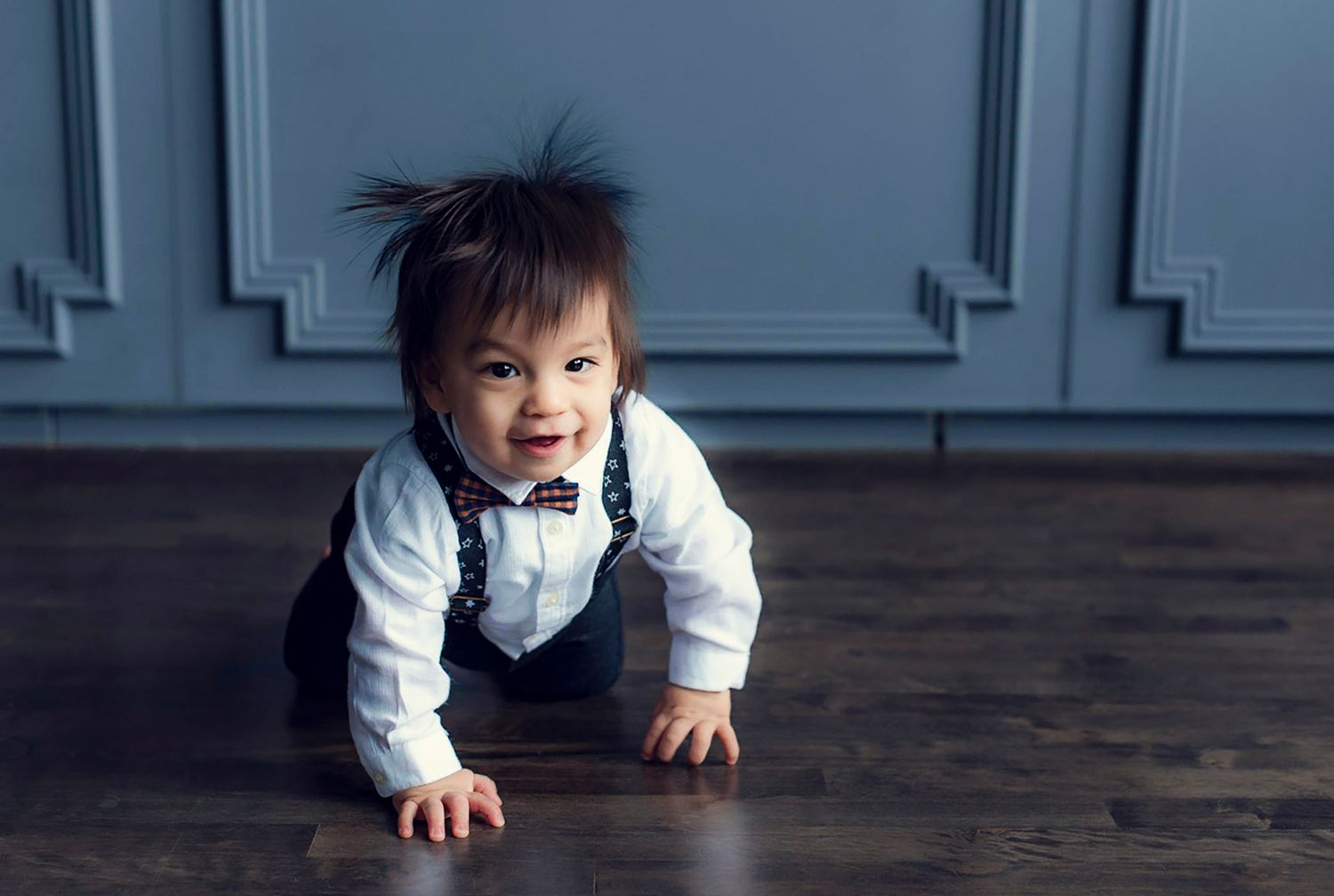 Happy Asian baby in suspenders and bow tie crawling on hardwood floor indoors.