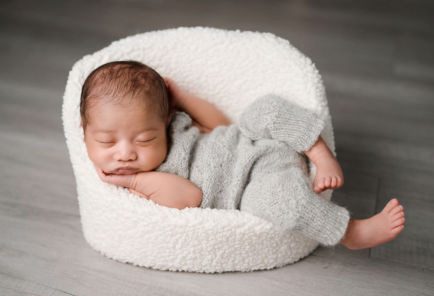 Sleeping newborn baby in a gray knit romper curled up in a cozy white boucle chair on a gray wood floor during a newborn photo session.