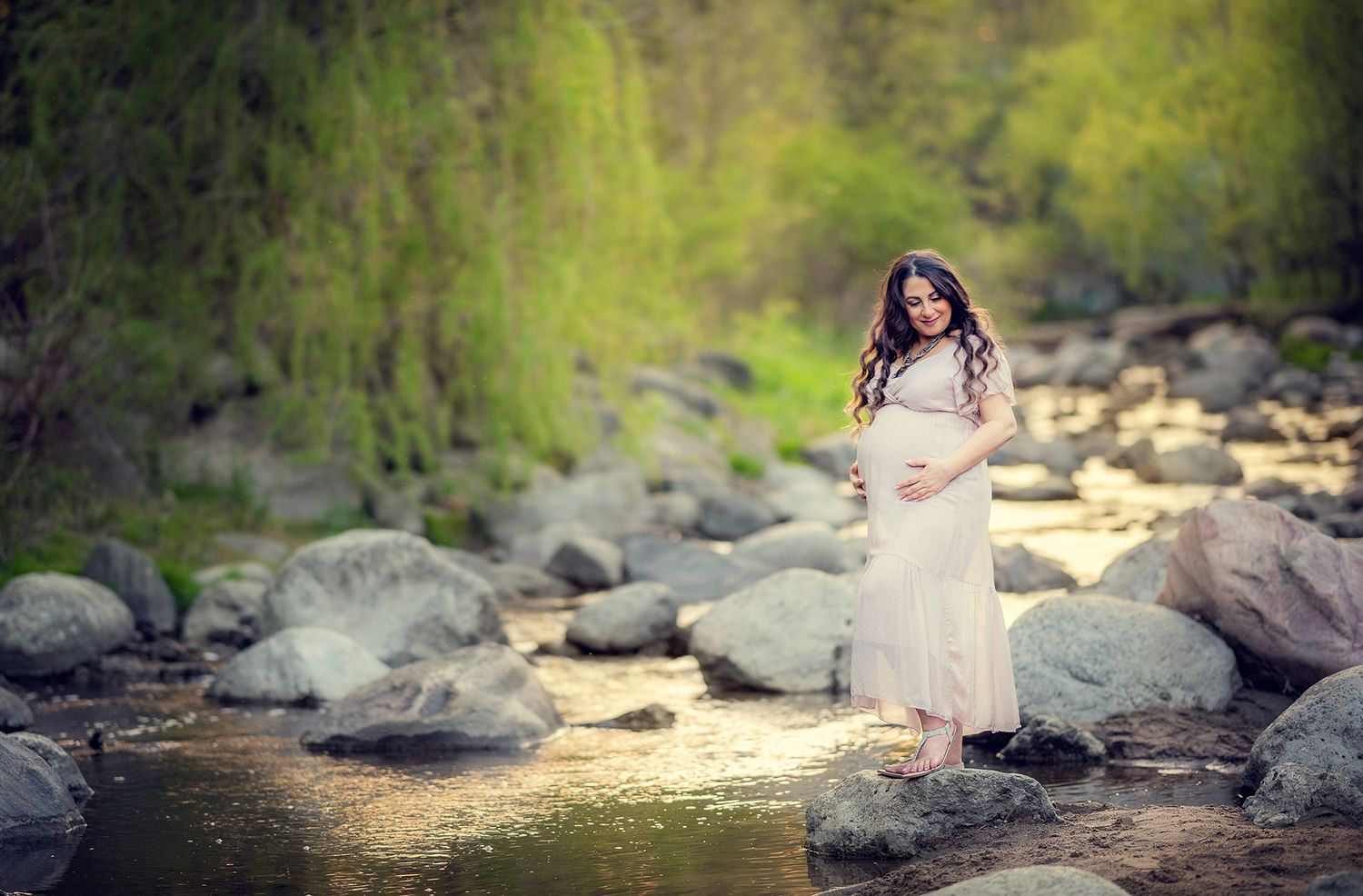 Pregnant woman in white dress standing on rocky riverbank surrounded by lush green trees.