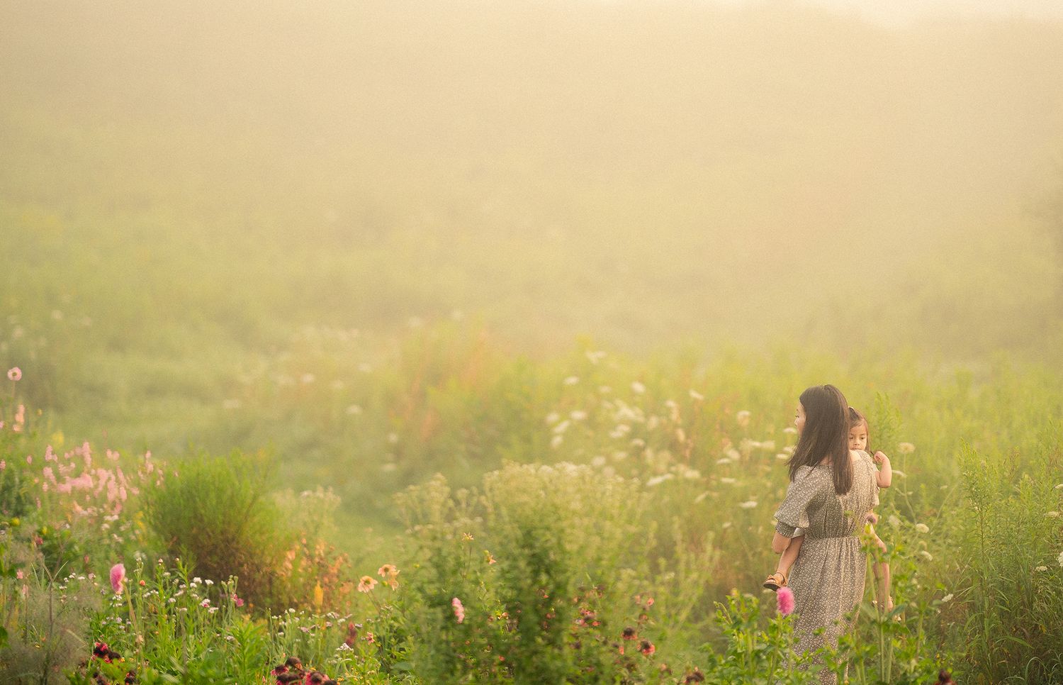 Woman in floral dress standing alone in a dreamy sunlit wildflower meadow with soft fog.
