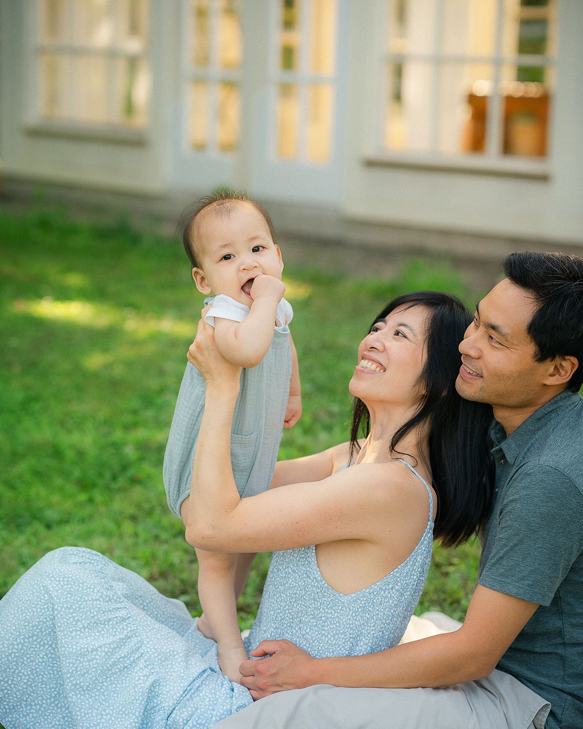 A happy Asian family sits on green grass, parents smiling as they lift their adorable baby outdoors near a house.