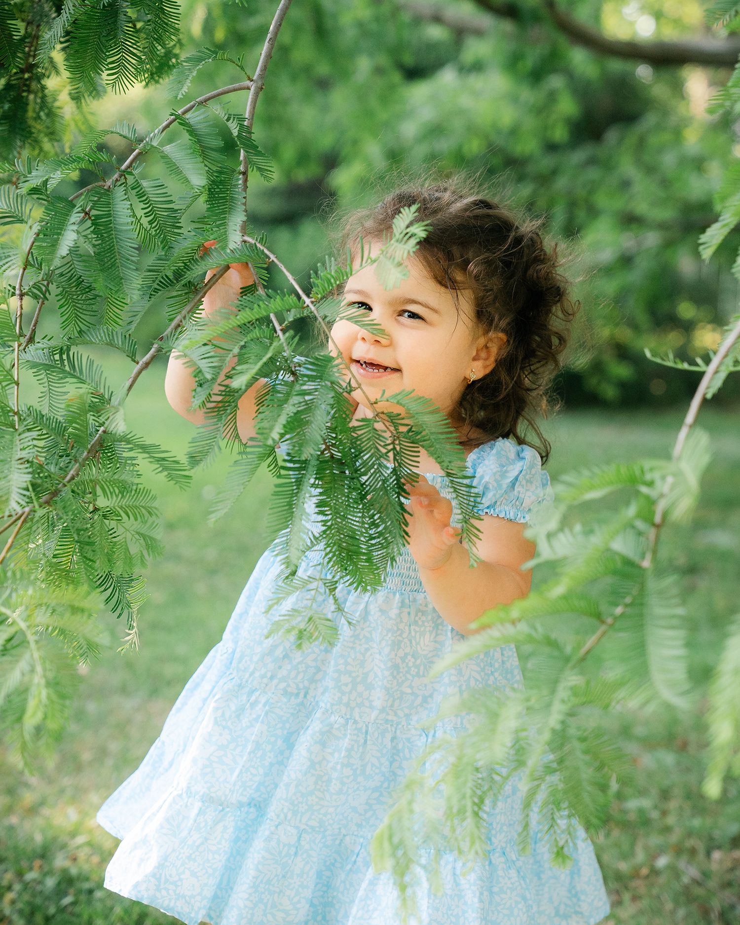 Smiling toddler girl in light blue dress playing with green tree branches outdoors.
