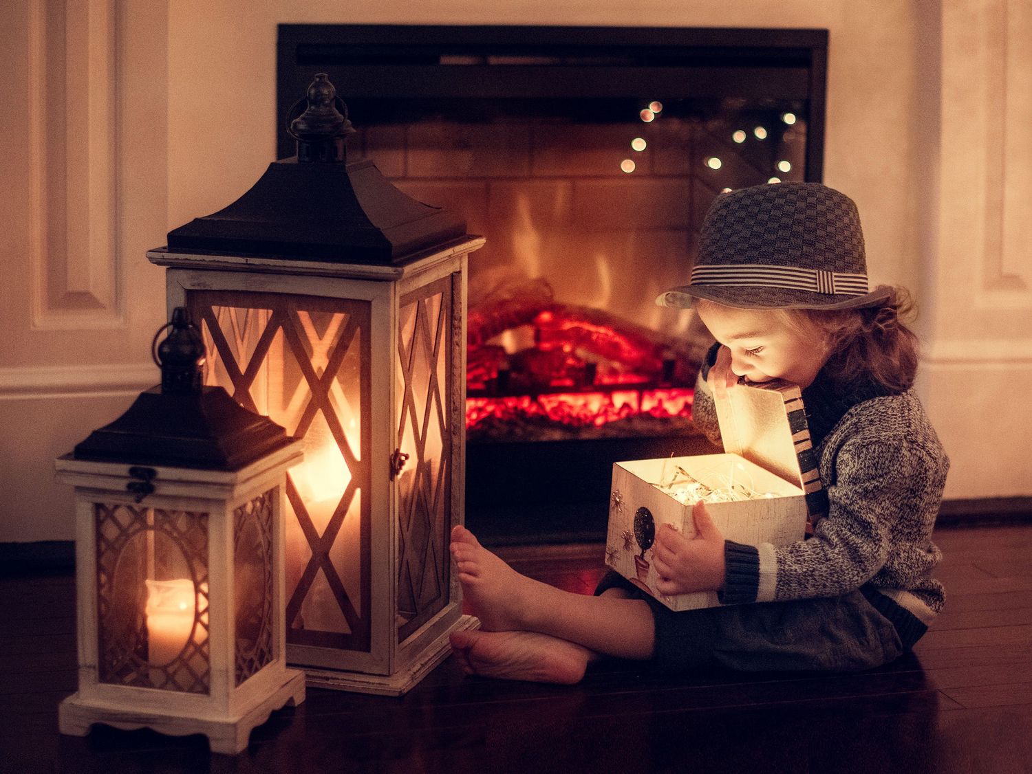 A child in a hat sits on the floor by glowing lanterns and a fireplace, opening a magical glowing gift box.