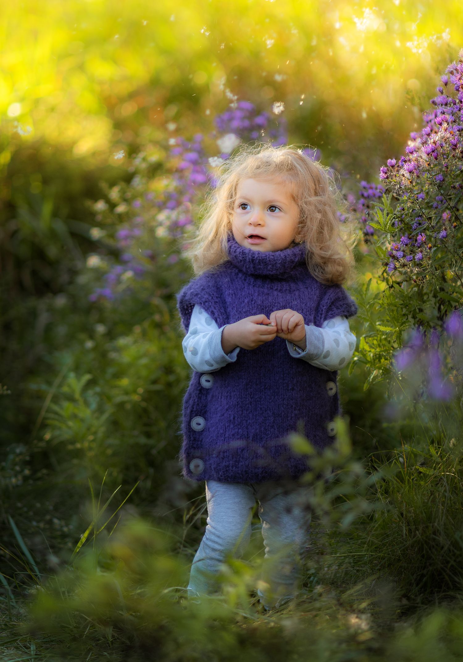 Cute blonde toddler girl in purple knit vest standing among wildflowers in sunny garden meadow.