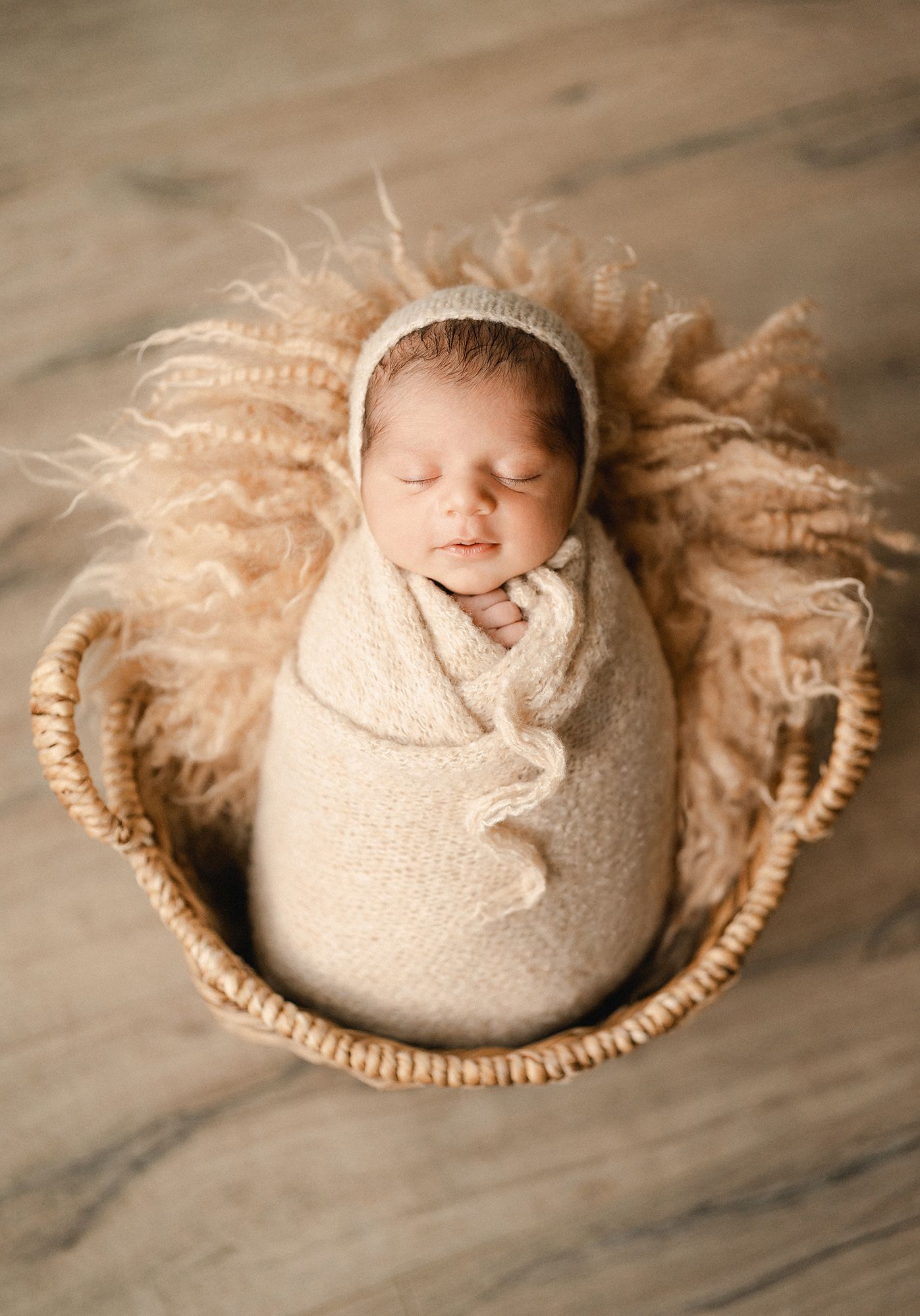 Sleeping newborn baby wrapped in a cream knit swaddle and bonnet, resting in a woven basket lined with fluffy beige fur.