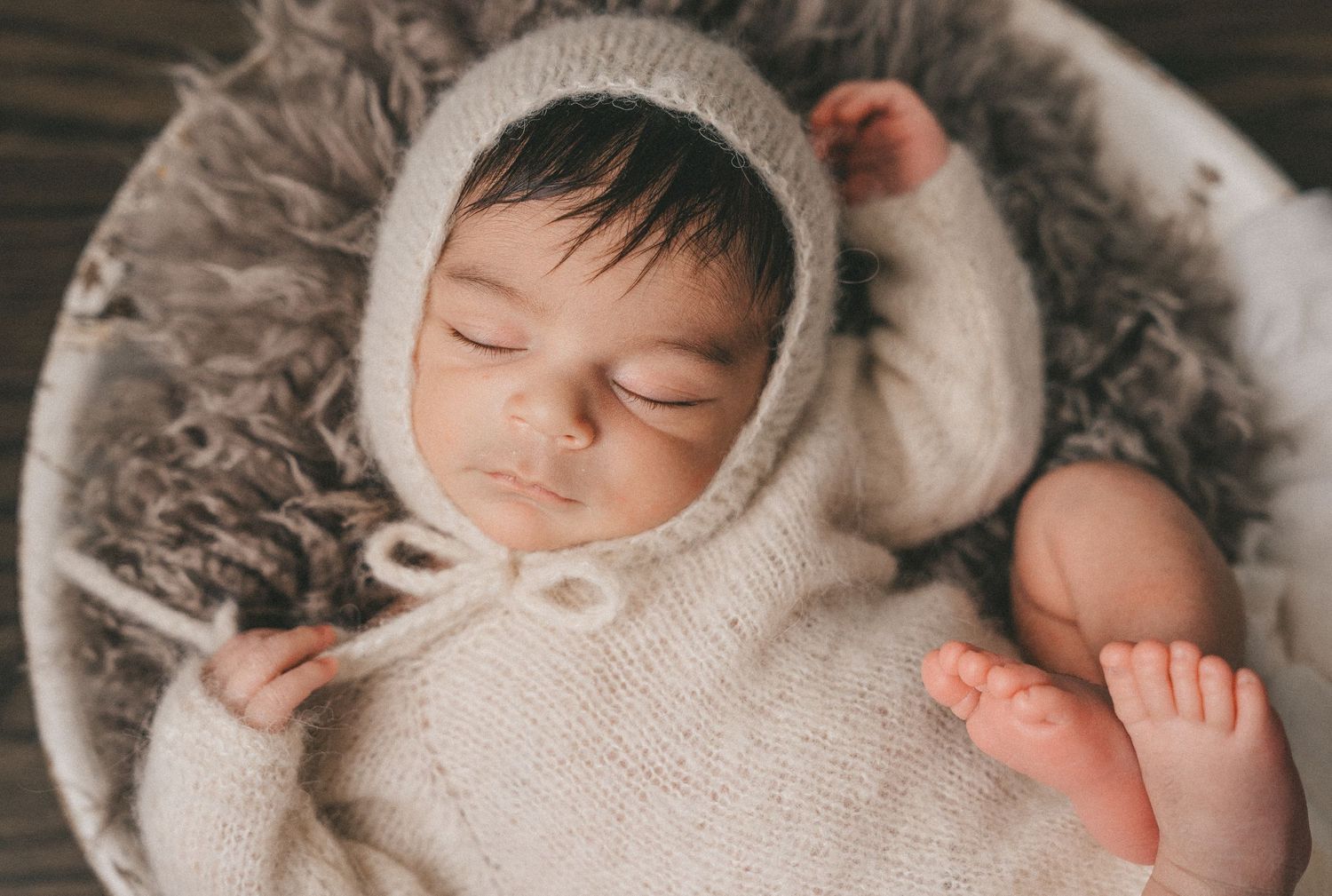 Sleeping newborn in a cream knit bonnet and romper posed in a round bowl with gray fur.